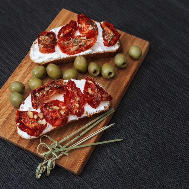 Bruschetta with sun-dried tomatoes and soft cheese, with green olives on a wooden board.