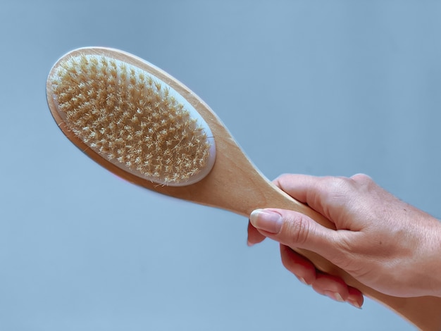 Brush with a handle for dry anti-cellulite massage or brushing in the hand of a girl on blue background.  Beauty concept.