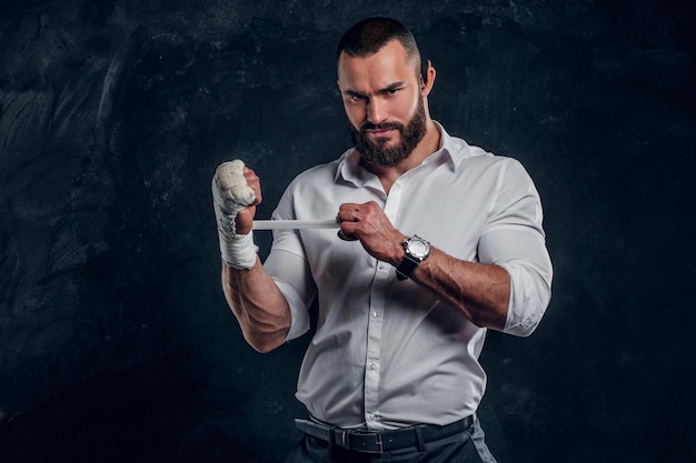 Brutal angry man in white shirt is wearing protection on his fist before fight at dark studio.