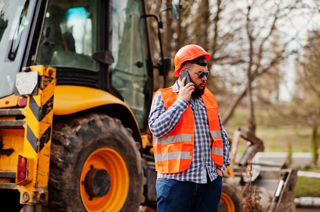 Brutal beard worker man suit construction worker in safety orange helmet, sunglasses against traktor with mobile phone at hand.