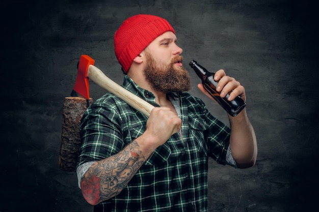 Brutal bearded lumberjack wearing green plaid shirt and red hat, drinking beer from a bottle and holds axe on a shoulder.