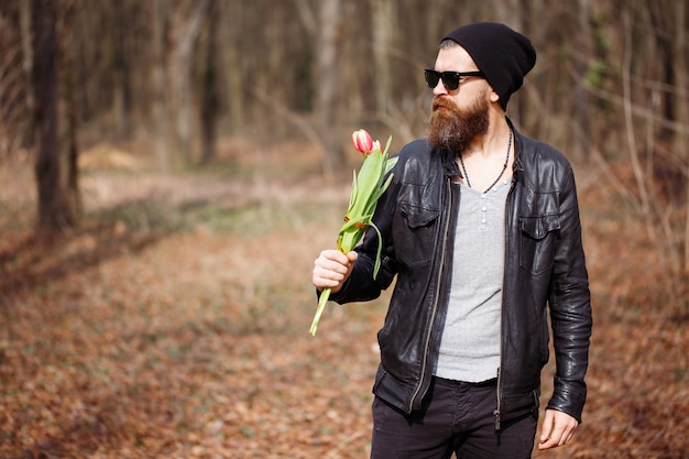 A brutal bearded macho man in a black leather jacket with gray hair in a warm hat and dark sunglasses holds a bouquet of red tulips