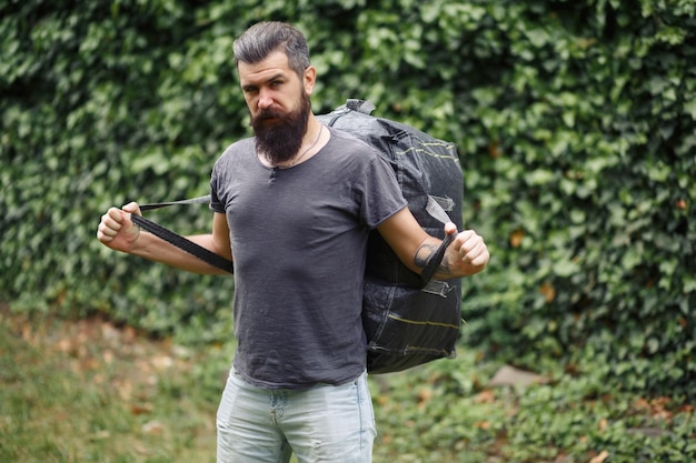 Brutal bearded male courier in shorts and a Tshirt with a big gray bag on his shoulders in a green park homeless not shaved man with a big gray bag