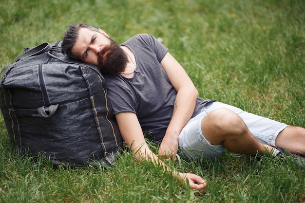 Brutal bearded male courier in shorts and a Tshirt with a big gray bag is resting in a green park homeless man not shaved with things in nature