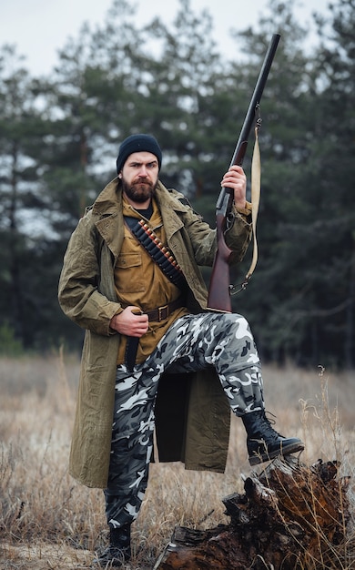 A brutal bearded male poacher in a black hat and khaki jacket and in a long cloak holds a gun, a large hunting knife and put his foot on a stump