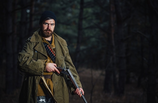 A brutal bearded man hunter in a dark hat and khaki jacket in a long cloak holds an unloaded gun against the background of a dark forest