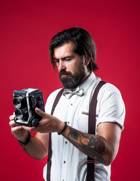 Brutal bearded man in suspenders holding retro camera while standing against red background photography