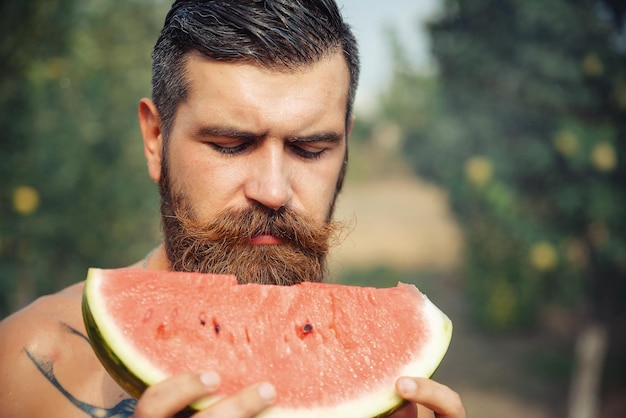 Brutal bearded man with a large red mustache and wet hair holds a slice of ripe red watermelon with his hands, a close-up portrait against the background of a green garden