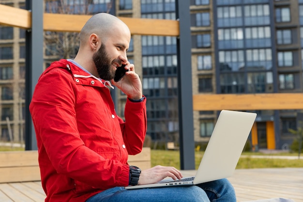 A brutal guy with a beard in everyday clothes works at a computer and talks on the phone on the street. Concept of working outside the home