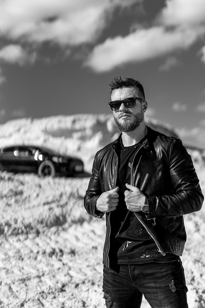 Brutal man standing on the road with luxury car background. Fashion model guy in stylish clothes. Black and white photo.