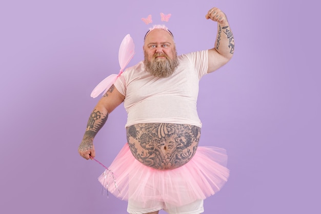 Brutal obese man with tattoos in fairy costume shows muscles on purple background