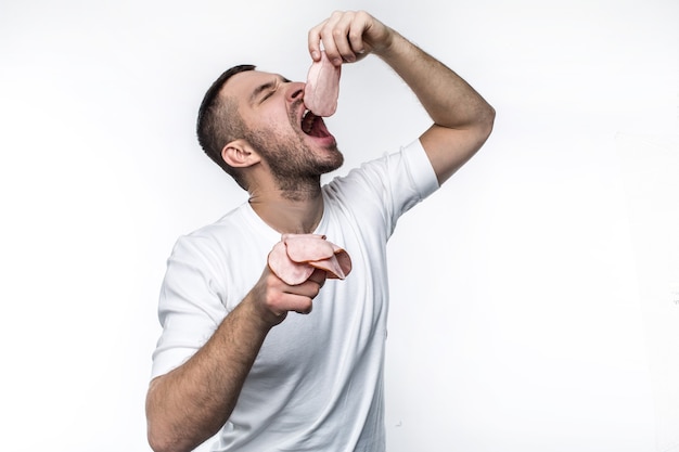 Brutal and real man is eating sliced pieces of meat. He is putting one piece of meat in the mouth and keeping the other pieces in the other hand. Isolated on white background