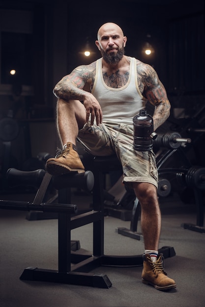 Brutal tattooed bearded man in the gym drinking water after workout. Fitness and bodybuilding. Caucasian man doing exercises in gym.