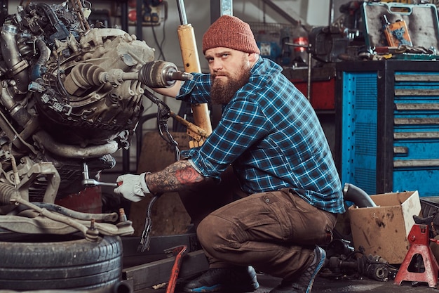 Brutal tattooed bearded mechanic specialist repairs the car engine which is raised on the hydraulic lift in the garage. Service station.