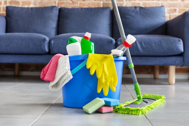 Bucket with sponges bottles of detergent and mopping stick in front of sofa in living room Housework cleaning equipment Cleaning service idea