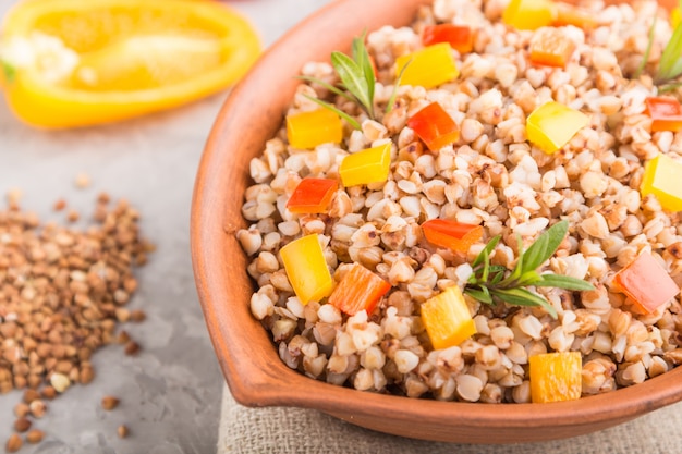 Buckwheat porridge with vegetables in clay bowl on a gray concrete surface and linen textile. Side view, close up, selective focus.