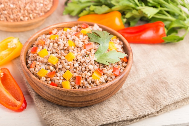 Buckwheat porridge with vegetables in wooden bowl on a white wooden surface and linen textile. Side view, close up, selective focus, macro.