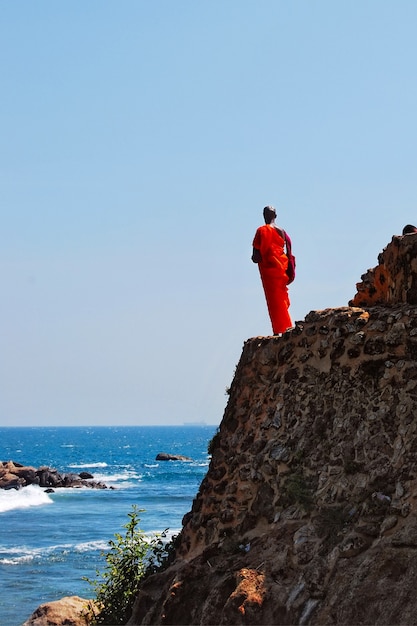 Buddhist monk stands on the rock and watches at sea, a ship goes there, Sri Lanka