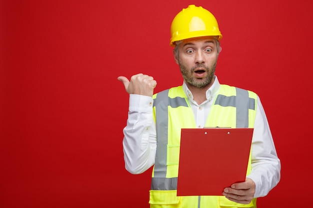 Builder man in construction uniform and safety helmet holding clipboard looking at it being surprised pointing with thumb to the side standing over red background