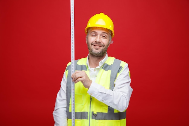 Builder man in construction uniform and safety helmet holding measure tape looking at camera smiling cheerfully happy and positive standing over red background