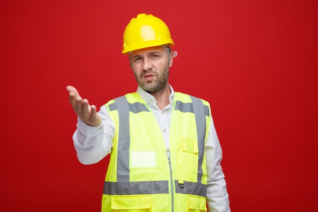 Builder man in construction uniform and safety helmet looking at camera confused and displeased raising arm in displeasure standing over red background