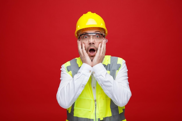 Builder man in construction uniform and safety helmet wearing safety glasses looking at camera amazed and surprised holding hands on hic cheeks standing over red background