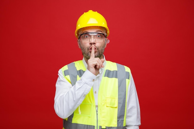 Builder man in construction uniform and safety helmet wearing safety glasses looking at camera making silence gesture with finger on lips standing over red background