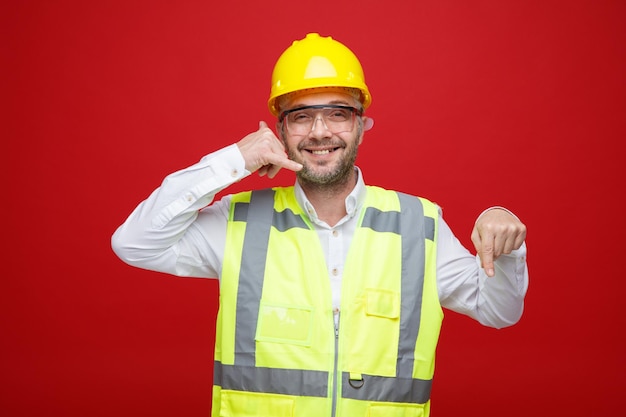 Builder man in construction uniform and safety helmet wearing safety glasses looking at camera smiling cheerfully making call me gesture pointing with index finger down standing over red background