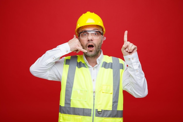 Builder man in construction uniform and safety helmet wearing safety glasses looking at camera surprised making call me gesture pointing with index finger up standing over red background