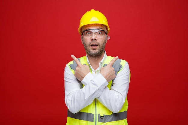 Builder man in construction uniform and safety helmet wearing safety glasses looking at camera surprised pointing with index fingers to opposite sides standing over red background