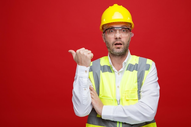 Builder man in construction uniform and safety helmet wearing safety glasses looking at camera surprised pointing with thumb to the side standing over pink background