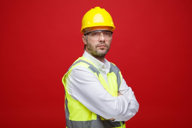 Builder man in construction uniform and safety helmet wearing safety glasses looking at camera with serious face with arms crossed on his chest standing over pink background