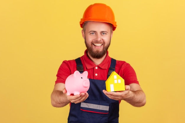 Builder man wearing blue uniform and protective helmet showing piggy bank and paper house