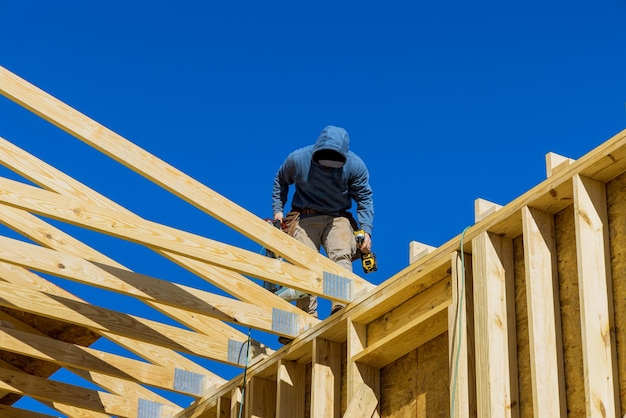 The builder on the unfinished roof with nailing wooden beams in frame house under construction