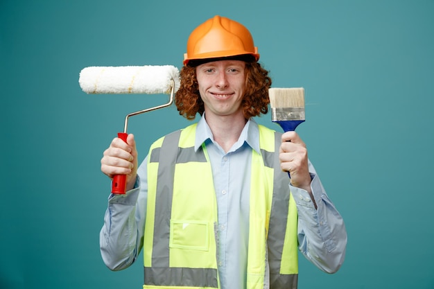 Builder young man in construction uniform and safety helmet holding paint roller and brush looking at camera happy and positive smiling standing over blue background