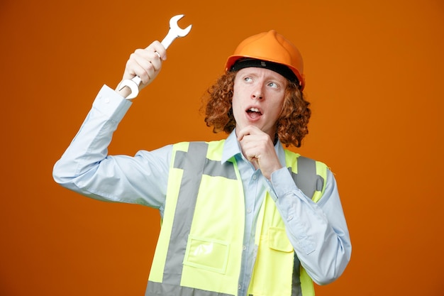 Builder young man in construction uniform and safety helmet holding wrench looking up with pensive expression thinking standing over orange background