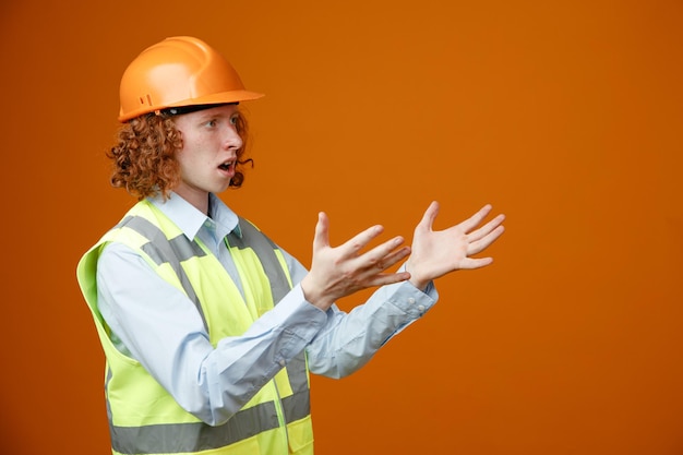 Builder young man in construction uniform and safety helmet looking aside being confused raising arms in displeasure standing over orange background