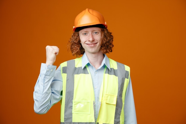 Builder young man in construction uniform and safety helmet looking at camera happy and positive smiling pointing with thumb back standing over orange background