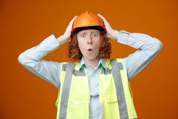 Builder young man in construction uniform and safety helmet looking at camera surprised holding hands on his head standing over orange background