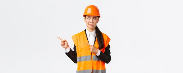 Building construction and industrial concept Indecisive asian female architect being unsure wearing reflective clothing and helmer smirk as pointing and looking upper left corner puzzled