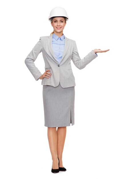 building, developing, advertising and architecture concept - friendly young smiling businesswoman in white helmet holding something on palm