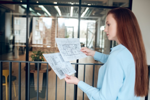 Photo building feature. smart young adult good looking woman with long red hair pondering over building plan standing indoors in front of glass wall