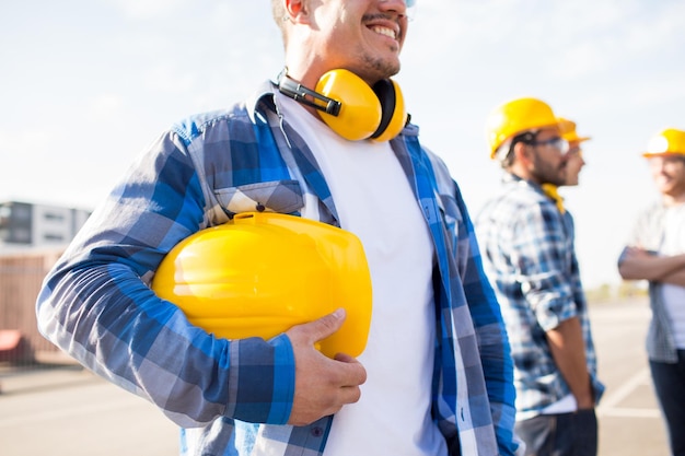 building, protective gear and people concept - close up of builder holding yellow hardhat or helmet at construction site