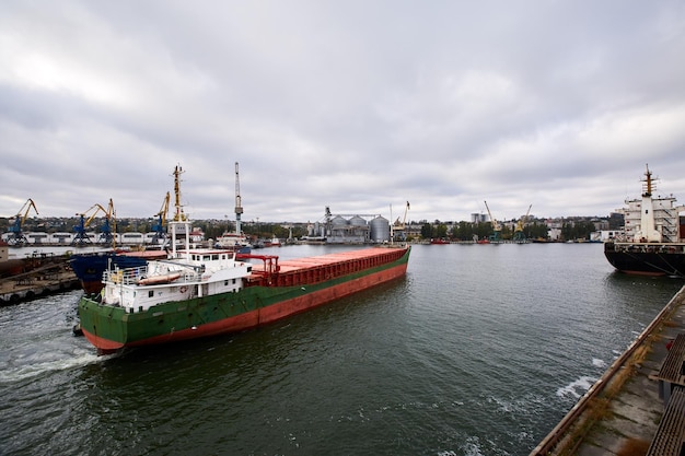 Bulk ship enters harbour of commercial dock at seaport to load at grain terminal wheat elevators