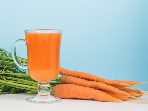 A bunch of carrots and a glass of smoothies on a white table on a blue background.