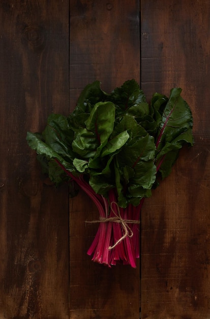 A bunch of green leaves of fresh swiss chard with red stalks tied with string on dark wood table V