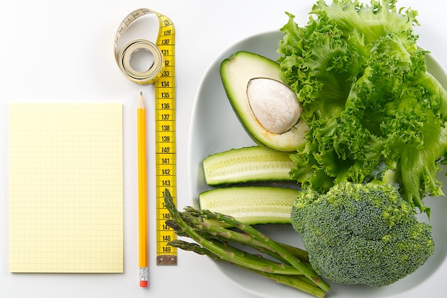 Bunch of vegetables laying on the plate, and near it is placed ruler, pen, and a note, and all of them are laying on the white background.