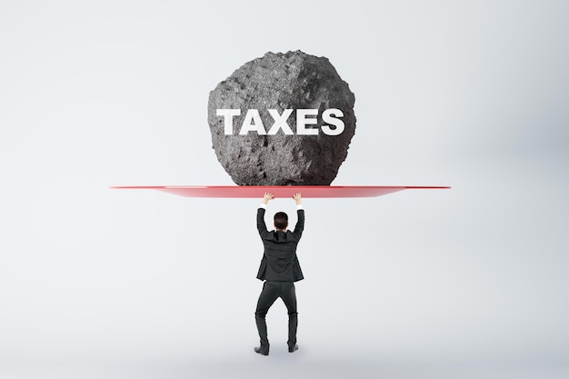 Burden of tax payments concept with businessman back view prevents huge stone from falling on his head with taxes sign on light background