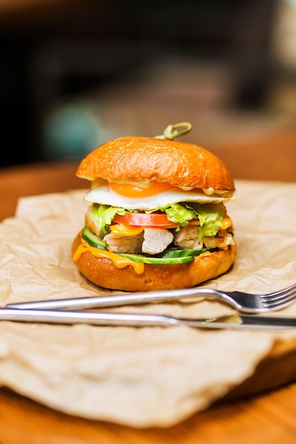 Burger with chicken pieces fresh cucumbers and tomatoes fried egg and cheese sauce on parchment paper on a wooden plate