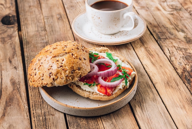 Burger with smoked salmon and cream cheese on plate and mug of coffee over rustic wooden background. Selective focus